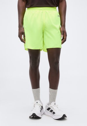 Άτομο που φοράει αθλητικό σορτς νέον κίτρινου χρώματος, γκρι κάλτσες και λευκά αθλητικά παπούτσια Adidas, στεκόμενο με τα πόδια ανοιχτά σε μια απλή φόντο.