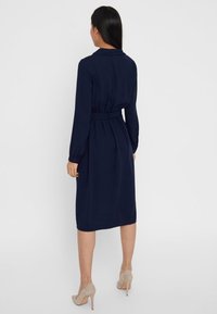 Abito blu navy con maniche lunghe, vita segnate da una cintura e orlo a lunghezza ginocchio. Tessuto liscio e design classico del colletto. Indossato con tacchi nude.