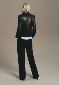 Blazer de piel negra con un diseño ajustado, que presenta una textura suave y un brillo sutil. Combinado con pantalones negros de pierna ancha y una camisa blanca.