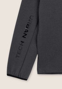 Maglietta grigia a maniche lunghe con un design aderente. Presenta testo nero lucido "TECH" e "URBAN" lungo la manica. Tessuto morbido.
