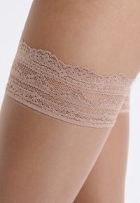 Nude pończochy sięgające za uda z koronkową górą w kwiatowy wzór. Pończochy charakteryzują się gładką fakturą oraz wzmocnionym palcem.