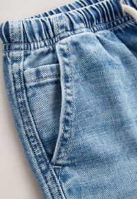 Lyseblå denim bukser med elastisk taljebånd og synlige syninger ved forlommerne, vist i nærbillede på en neutral baggrund.