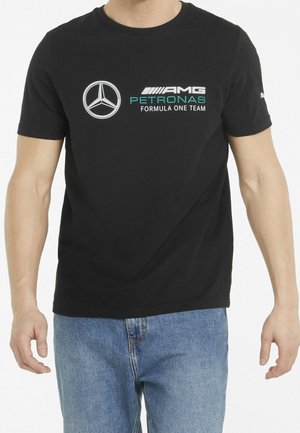 Mężczyzna w czarnej koszulce zespołu Mercedes AMG Petronas Formula One oraz niebieskich dżinsach, stojący na białym tle.