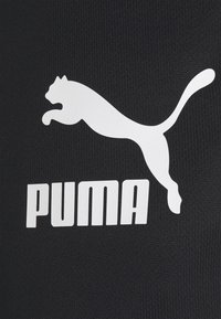 Melna auduma virsma ar lielu baltu Puma logotipu, kurā virs vārda "PUMA" treknajos baltajos burtos atrodas lecošas kaķa siluets.