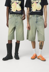 Deux individus portant des shorts en denim vert clair surdimensionnés. L'un porte des bottes noires jusqu'au genou ; l'autre arbore des baskets en daim orange avec des chaussettes blanches.