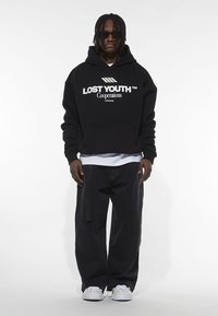 Hoodie cropped nero con grafica "LOST YOUTH", abbinato a pantaloni neri larghi e sneakers bianche. Gli accessori includono occhiali da sole.