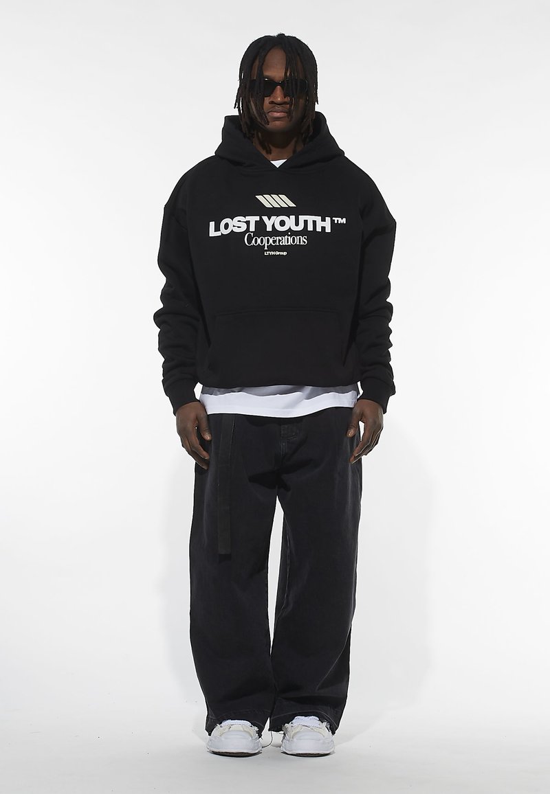 Hoodie cropped nero con grafica "LOST YOUTH", abbinato a pantaloni neri larghi e sneakers bianche. Gli accessori includono occhiali da sole.
