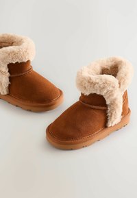 Brune ruskind ankelstøvler med en fluffy beige pelskant og foring. Sålens farve er tan gummi med et tekstureret grebsmønster.