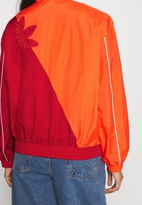 Person som bär en röd och orange jacka med färgblockering och en sydd trefoil-logga på vänster bakaxel samt blå jeans.