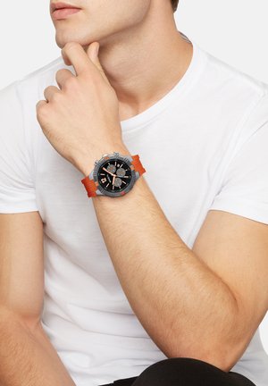 Mand i hvid skjorte hviler hagen på hånden iført sort og sølv chronografur med orange gummirem.