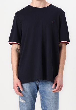 Mann trägt ein dunkelblaues kurzärmeliges T-Shirt mit roten, weißen und blauen Abschlüssen an den Ärmeln und hellblaue zerrissene Jeans, steht vor einem weißen Hintergrund.