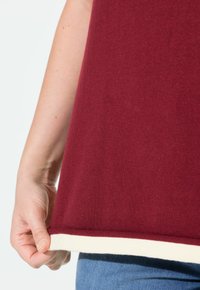 Hand die de zoom van een bordeaux top met een witte rand vasthoudt, gedragen over blauwe jeans, tegen een effen achtergrond.