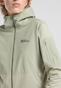 Hellgrüne Softshelljacke mit Reißverschluss vorne, ausgestattet mit einer Kapuze, seitlichem Taschen und einem Logo auf der Brust. Glatte Textur, sportliches Design.