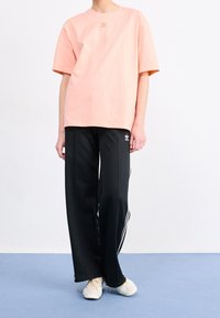 Rosa Baumwoll-T-Shirt mit Logo, kombiniert mit schwarzen Jogginghosen mit weißen Seitenstreifen. Das Modell trägt cremefarbene Sneakers.