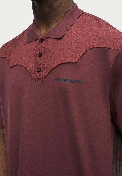 Άνδρας που φοράει πουκάμισο πόλο μαρόν με υφασμάτινη λοξή ραφή, τρία κουμπιά και την κεντημένη επιγραφή "LES BENJAMINS" στο στήθος.