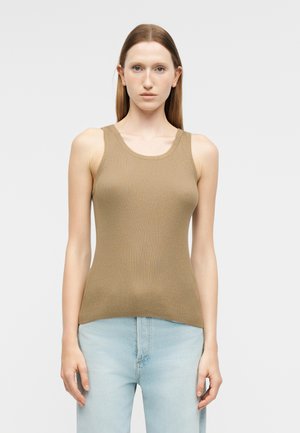 Kvinde med glat hår iført en beige ribstrikket, ærmeløs top og lyseblå jeans, stående foran en ensfarvet lys baggrund.