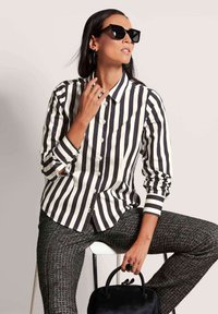 Zwart-wit gestreepte overhemden met lange mouwen, gecombineerd met gestructureerde donkere broek. Accessoires omvatten cat-eye zonnebril en een zwarte handtas.