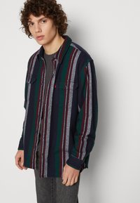 Camisa de franela para hombre con rayas verticales en azul marino, verde y burdeos, dos bolsillos en el pecho, cierre de botones y mangas largas. Textura suave.