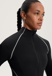 Fekete cipzáras sportdzseki fél gallérral, szürke díszítéssel a varrások mentén és testhez álló kialakítással a karcsú megjelenés érdekében.