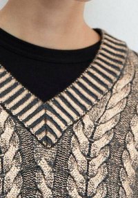 V-paita, jossa on metallinhohtoiset kultaiset ja mustat raidat kauluksessa sekä kultainen palmikkoneulekuvio mustan paidan päällä henkilön ylävartalolla.