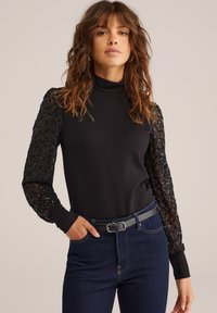 Melna turtlenecks blūze ar puscaurspīdīgām mežģīņu piedurknēm, kas rotātas ar dimantu rakstu un pērlītēm, saskaņota ar tumšajiem džinsiem un jostu.