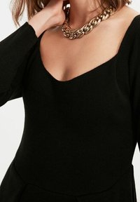 Black knitted sweater with a wide neckline, long sleeves, and smooth texture. Paired with a large gold chain necklace.