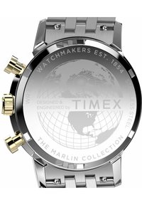 Reloj de la colección Timex Marlin con correa de acero inoxidable plateado, caja redonda, detalles en oro y un mapa mundial grabado en la parte posterior.