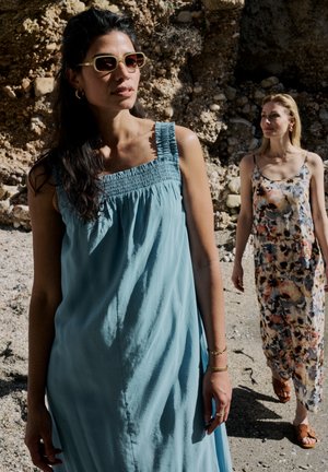 Deux femmes marchent près d’un terrain rocheux ; l’une porte une robe bleue sans manches avec des lunettes de soleil, l’autre une robe longue fleurie sans manches et des sandales.