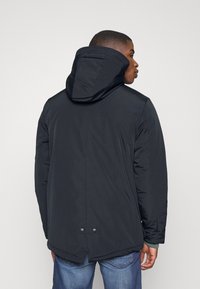 Svart isolert jakke med hette, med lengre bakside, trykknappdetaljer og glatt tekstur. Stylet med jeans.