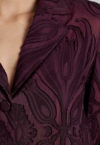 Donker bordeaux jacket met een getextureerd bloemendessin en een opvallende glans. Gestructureerde kraag en revers, met ingewikkelde details langs de randen.