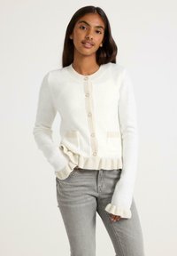 White cardigan with a round neckline, front pockets, and ruffled cuffs. Features beige accents and buttons, made from fluffy material.