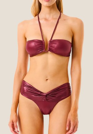Donna che indossa un bikini senza spalline lucido color amaranto con collo halter e fascia attorcigliata sulla parte inferiore, in piedi davanti a uno sfondo beige uniforme.