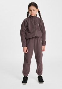 Fleece-Trainingsanzug in dunklem Mauve mit einer Jacke zum Reißverschluss und elastischen Bündchen. Besitzt ein kleines Logo auf der Brust und an den Hosen. Schwarze Sneakers.