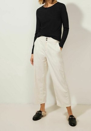 Person iført sort langærmet top, hvidt højtaljet bukser med vide ben og sorte slip-on loafers med beige pelsforing. Hænderne i lommerne.