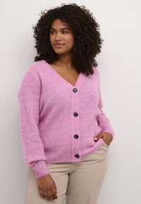 Cardigan roz cu un model texturat, guler în V, cinci butoane negre pe față și manșete în dungi. Purtați deasupra unor pantaloni bej.