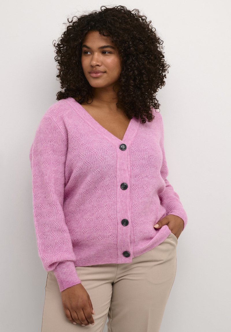 Cardigan roz cu un model texturat, guler în V, cinci butoane negre pe față și manșete în dungi. Purtați deasupra unor pantaloni bej.