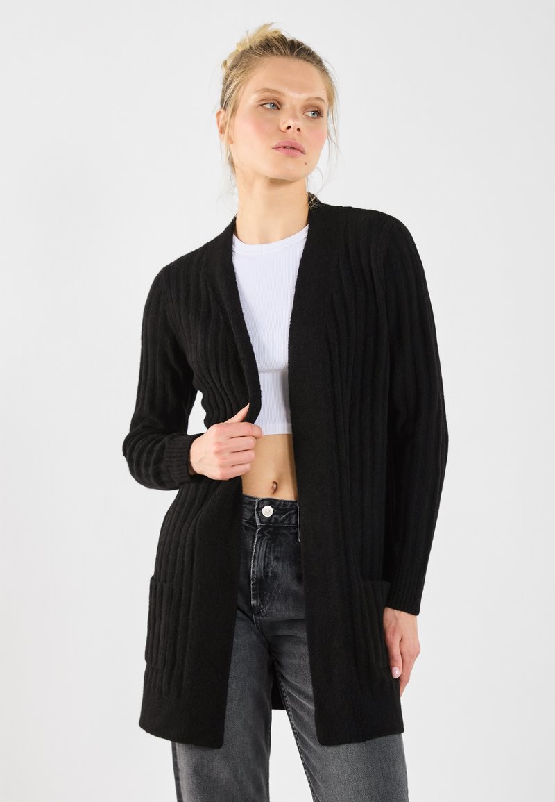 Zwarte geribbelde cardigan van zachte stof, met een open voorkant, lange mouwen en twee voorzakken. Gestyled met hoge taille jeans.