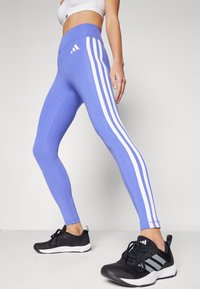 Leggings viola con triple strisce bianche, vita alta, design aderente, texture liscia. Abbinati a scarpe da ginnastica nere con tomaia in mesh.