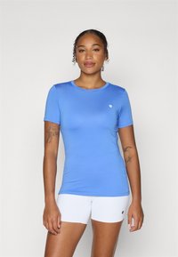 Camiseta azul justa de mangas curtas, com decote redondo e um pequeno logo branco. Usada com calções brancos, de textura suave e tecido leve.