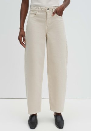 Persoon die beige broek met hoge taille en wijd uitlopende pijpen draagt, voorzien van voorzakken, en zwarte instapschoenen met vierkante neus, tegen een effen achtergrond.