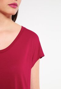 Kvinde iført en kortærmet, dyb pink top med en bred, rund halsudskæring mod en simpel hvid baggrund.