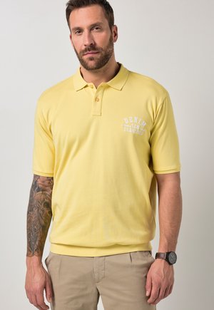 Homme aux cheveux courts et barbe porte un polo jaune avec le texte "Denim San Francisco", un pantalon beige, une montre-bracelet et une manche tatouée sur le bras gauche.