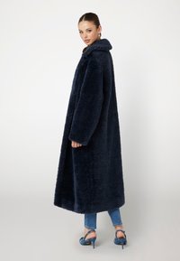 Marineblå kunstlæderfrakke med bred krave, lange ærmer og en afslappet pasform, der når til midt på læggen, kombineret med tætsiddende jeans og højhælede sko.