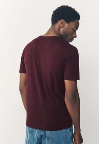 Burgundia lühikeste varrukatega puuvillane t-särk, millel on ümar kaelus ja lõõgastav istuvus, kantud koos siniste teksapükstega.