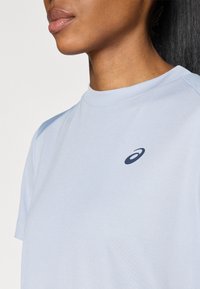 Ανοιχτό μπλε αθλητικό μπλουζάκι με στρογγυλή λαιμόκοψη, κοντά μανίκια και σκούρο μπλε λογότυπο στο στήθος. Λείο ύφασμα, χαλαρή εφαρμογή.