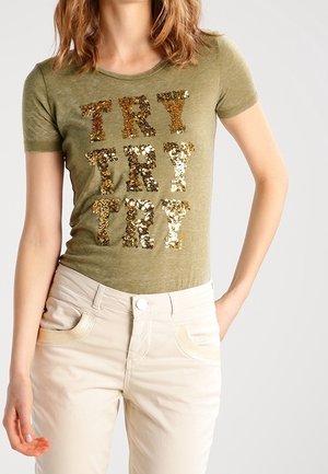 T-shirt verde oliva a maniche corte con testo in paillettes dorate "TRY" in lettere impilate. Abbinato a pantaloni beige chiaro con tasche decorative.