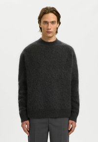 Homme aux yeux bleus et cheveux bruns mi-longs portant un pull gris foncé en tissu duveteux et un pantalon gris, debout devant un fond blanc.