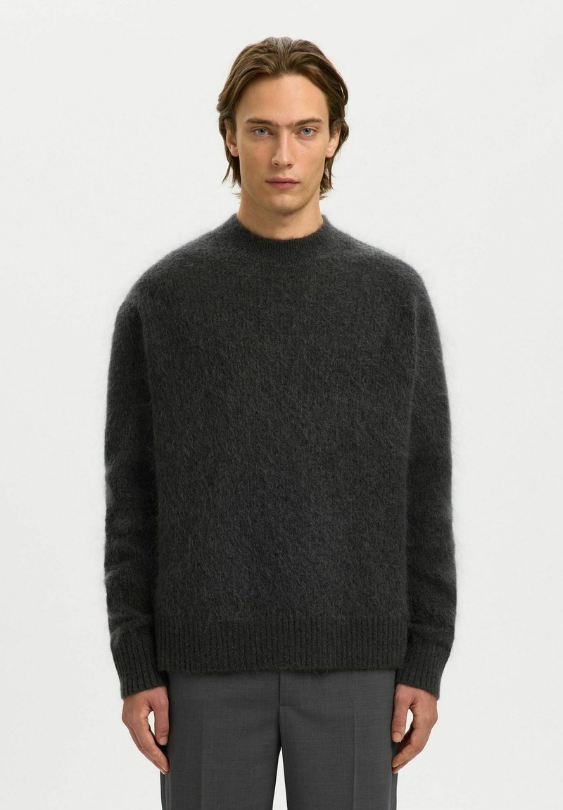 Homme aux yeux bleus et cheveux bruns mi-longs portant un pull gris foncé en tissu duveteux et un pantalon gris, debout devant un fond blanc.