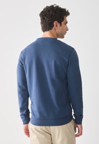 Mann med mørkt hår iført en ensfarget blå langermet skjorte og beige bukser, stående med ryggen mot en hvit bakgrunn.