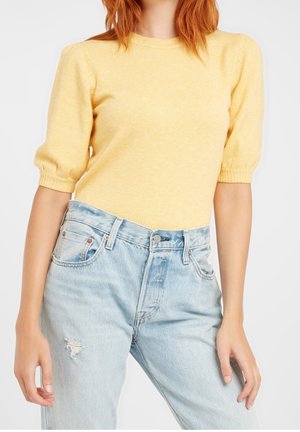 Femme portant un pull jaune à manches courtes et un jean déchiré bleu clair taille haute, debout devant un fond uni.